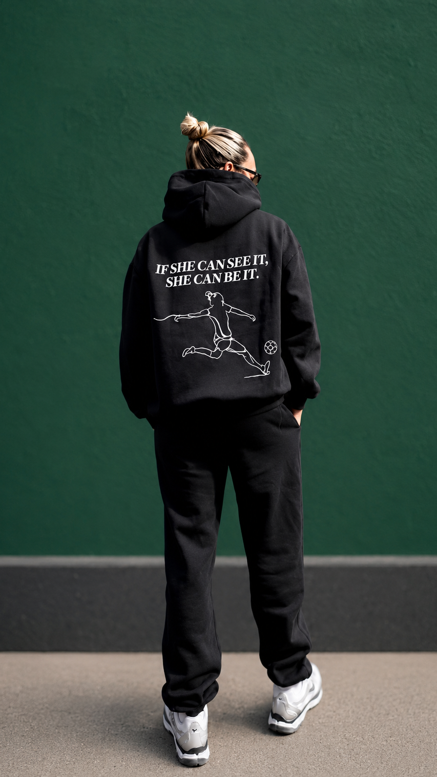 Person wearing a black hoodie with text and graphics on the back against a green wall.